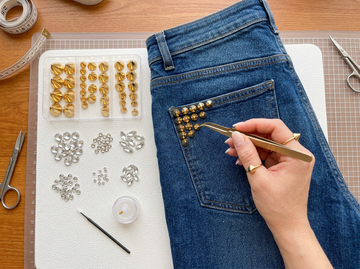 Person using tweezers to apply gold studs to the back pocket of blue jeans.