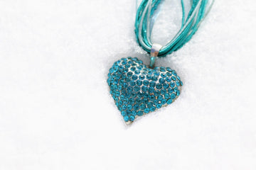 Blue rhinestone heart pendant necklace resting on white snow, with turquoise cords and shimmering crystal accents.
