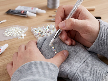 Hands applying iridescent rhinestones to a gray hoodie using a crystal pickup tool.