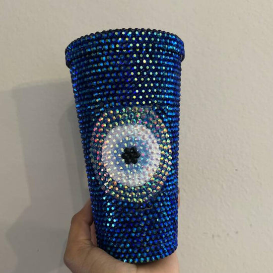 A travel cup covered in dark blue rhinestones, with an eye created from rhinestones on the front.